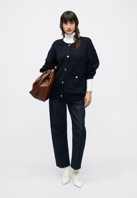 Tumesinine oversize-kardigan kuldsete nuppudega, tekstuurse kangaga, ühendatud valge poolkaeluse, tumedate teksade ja valgete teravate saapadega. Pruun käekott.