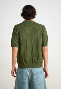 Strikket grøn genser med korte ærmer, som har et strukturert rutemønster, rund krage og en tettsittende midje. Båret med lyseblå denimshorts.