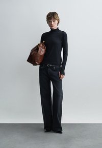 Γυναίκα με μαύρο πουλόβερ ζιβάγκο και φαρδιά μαύρα τζιν κρατά μεγάλη καφέ δερμάτινη τσάντα μπροστά σε απλό γκρι φόντο.