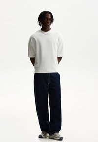 Bijela majica kratkih rukava, labavi tamnoplavi traperice s vidljivim šavovima, i svijetlosivi tenisi s crnim naglascima.