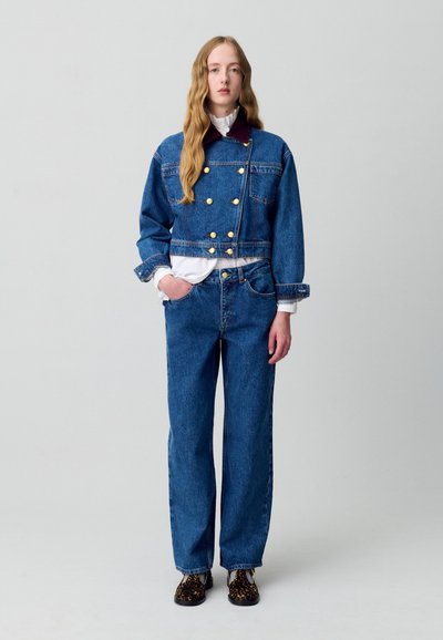 Giacca di jeans con bottoni dorati, design cropped e tonalità blu, abbinata a pantaloni in denim blu a vestibilità ampia e scarpe con stampa leopardata nera.