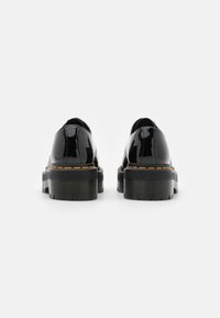 Scarpe in pelle verniciata nera con suole in gomma spessa e seghettata e cuciture gialle attorno alla piattaforma. Punta arrotondata e tacco basso.