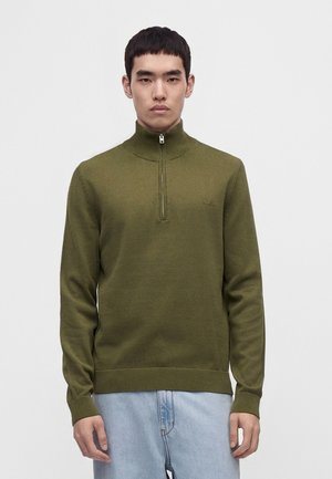 Jeune homme portant un pull vert olive à demi-fermeture éclair et un jean bleu clair, debout devant un fond blanc uni.