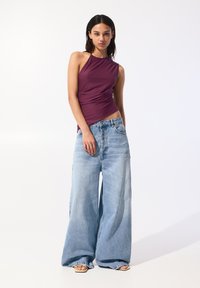 Haut sans manches violet avec un design plissé, associé à un jean en denim bleu clair à jambes larges. Le modèle se tient contre un fond blanc uni.