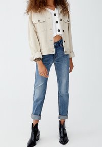 Mujer vestida con una chaqueta de pana beige, blusa blanca de botón recortada, jeans azules con puños y botines de tacón negros sobre un fondo blanco.
