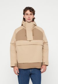 Veste à capuche beige avec un col haut, design bicolore, poches latérales et détails de poignets côtelés, portée sur un jean bleu.