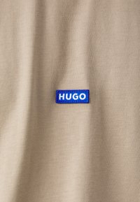 T-shirt en tissu khaki avec un patch rectangulaire brodé bleu portant le mot "HUGO" en lettres blanches. Texture lisse et design simple.