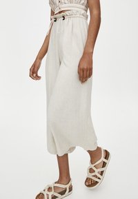 Pantalons culottes en lin beige avec taille à cordon et ourlet en forme de vagues, associés à des sandales tressées beige avec un design de lanières croisées.