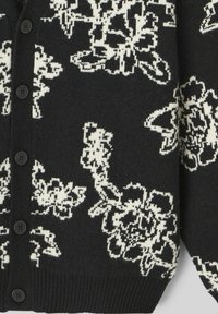 Cardigan noir avec motif floral blanc, texture tricotée, cinq boutons noirs et ourlet côtelé.