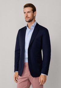 Blazer blu navy con due tasche frontali, chiusura a un bottone, indossato sopra una camicia azzurra chiara, abbinato a pantaloni rosa chiaro. Tessuto liscio.