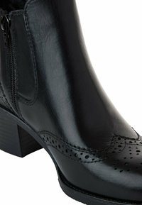 Bottine en cuir noir avec détails brogue, panneau élastique latéral, fermeture éclair et talon bloc empilé.