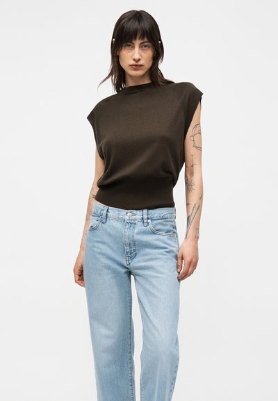 Γυναίκα με σκουρόχρωμα μαλλιά φορέσει αμάνικη σκούρα καφέ μπλούζα και ανοιχτόχρωμο μπλε τζιν, στέκεται μπροστά σε απλή ανοιχτή επιφάνεια.