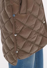 Brun vadderad pufferjacka med ett diamantmönster, sidofickor och tryckknappsdetaljer. Har en rundad nederkant och slät textur.