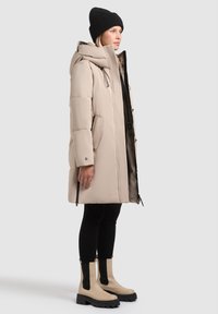 Manteau en duvet beige avec capuche, doublure noire, poches zippées, porté avec des leggings noirs et des bottines beiges à semelles épaisses.