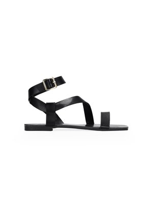 Sandale plate en cuir noir avec une bride de cheville, des brides croisées sur le pied et un design minimaliste. Elle présente un bout carré et une semelle rectangulaire.