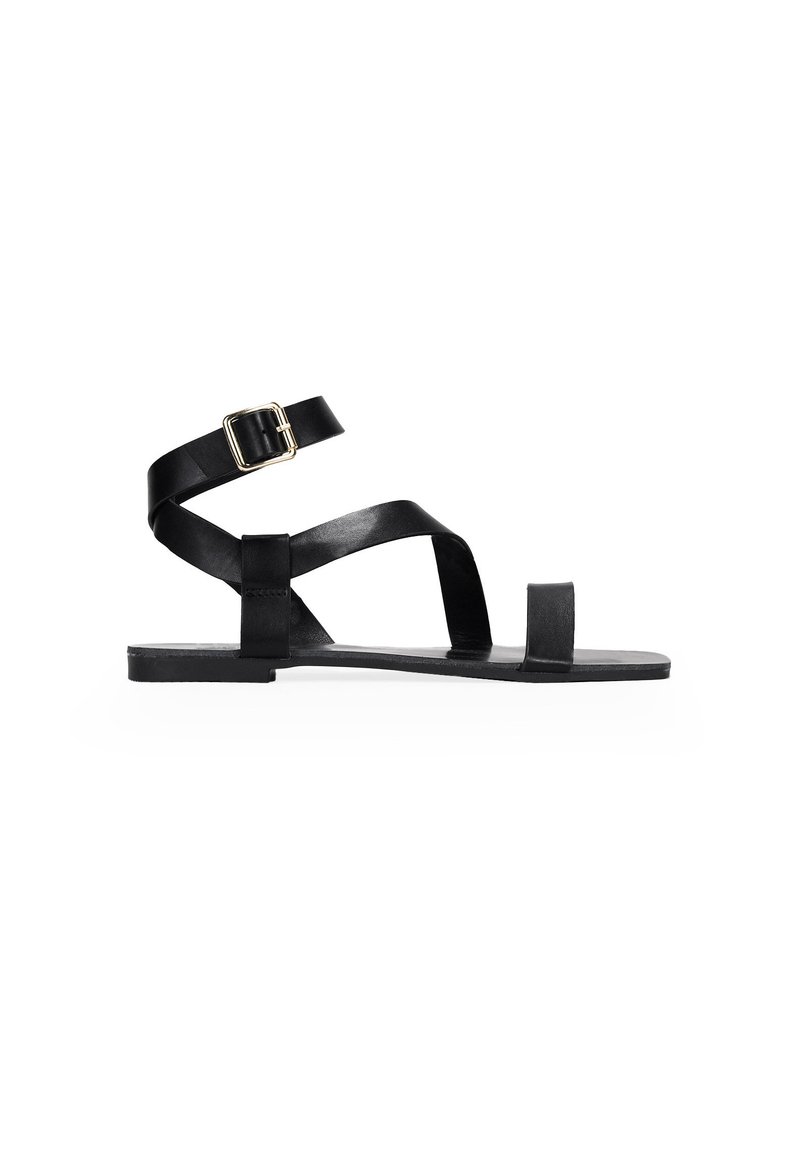 Sandal in pelle nera flat con cinturino alla caviglia, cinturini incrociati sopra il piede e design minimalista. Presenta una punta quadrata e una suola rettangolare.
