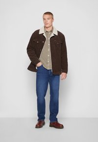 Chaqueta de pana marrón con cuello de borreguito, camisa beige de botones, jeans azules y botas de cuero marrón, sobre un fondo blanco.