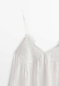 Haut blanc sans manches avec fines bretelles et broderie en relief complexe sur la partie supérieure de la poitrine, sur un fond uni.