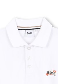Witte poloshirt met een klassieke kraag, drieknoopsluiting en een logo aan de linkerkant met rode en zwarte accenten. Gestructureerde stof.