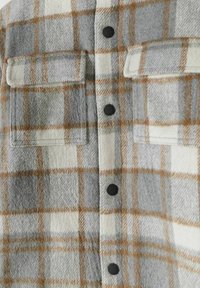 Chemise en flanelle grise et blanche avec un motif à carreaux marron, dotée de deux poches poitrine et de boutons noirs. Texture douce, design à carreaux.