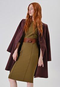 Olivengroene aansluitende jurk met naden als detail, gecombineerd met een kastanjebruine mantelcape en een bruine riem met een strikaccent. Gladde textuur.