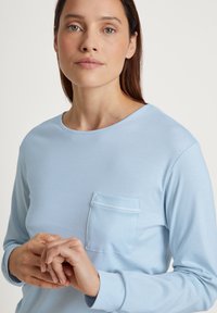 Lichtblauw langarm t-shirt van zacht materiaal. Ronde halslijn, borstzak en een ontspannende pasvorm met subtiele textuur en stikseldetails.