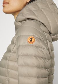 Gevoerde jas in lichtbeige met horizontale stiksels. Voorzien van een trekkoord capuchon en een oranje cirkelvormig logo op de bovenarm.