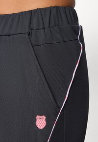Črne športne hlače z elastičnim pasom, subtilno teksturo, roza stranskimi akcenti in roza K-Swiss logotipom na prednjem žepu.
