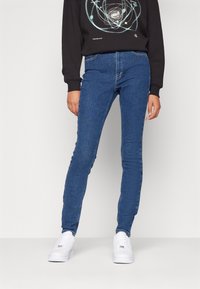 Tunna blå denimjeans med en slät textur, hög midja och klassisk femfickdesign, i kombination med vita sneakers och en svart sweatshirt.