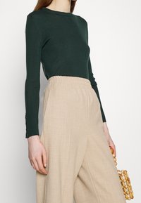 Kobieta ubrana w dopasowaną ciemnozieloną bluzk�ę z długim rękawem i luźne beżowe spodnie z wysokim stanem, trzymająca małą torebkę z drewnianych koralików.