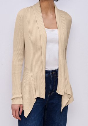Personne portant un cardigan beige côtelé ouvert sur l'avant, par-dessus un haut blanc et un jean bleu foncé, visible du cou jusqu'aux hanches sur un fond uni.