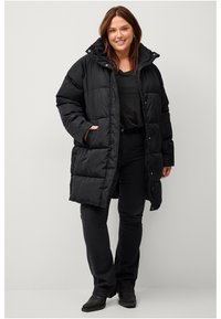 Sort puffet jakke med quiltet design, hætte og trykknapper. Har sidelommer og en afslappet pasform. Matchet med en sort top og jeans.