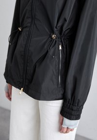 Svart zip-up jacka med elastiska muddar och justerbara dragskor, bärs över vita byxor och ljusblå fingerlösa handskar.