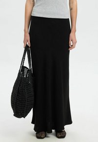 Jupe maxi noire en tissu doux et fluide, assortie à un sac tissé noir texturé et des chaussures en maille à bout ouvert. Design et silhouette simples.