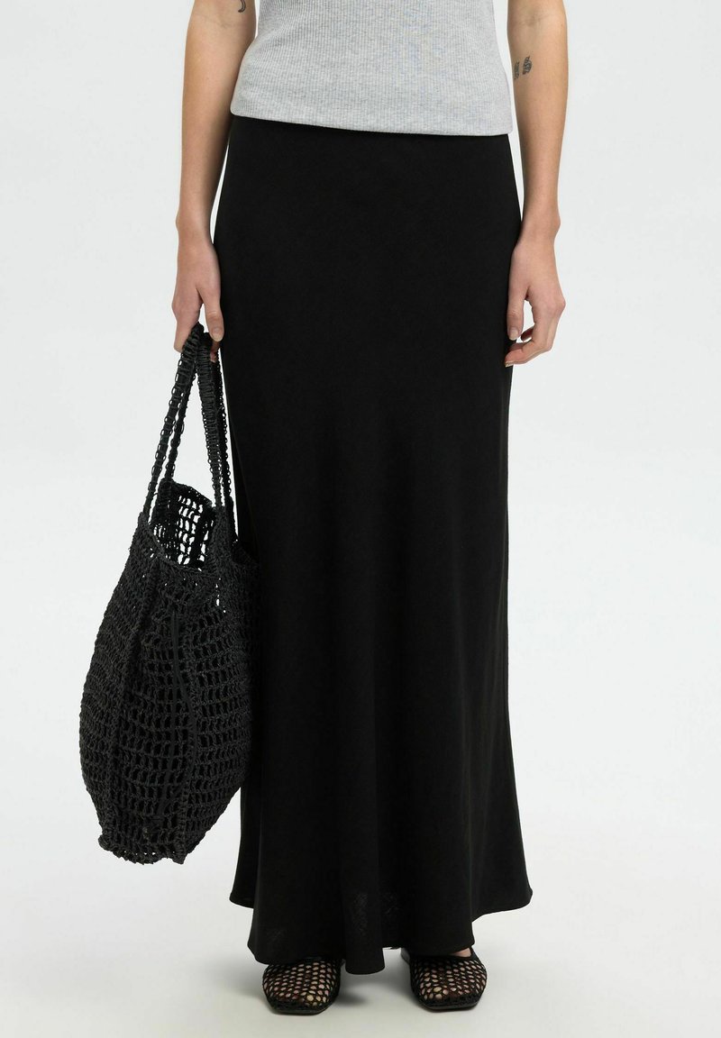 Jupe maxi noire en tissu doux et fluide, assortie à un sac tissé noir texturé et des chaussures en maille à bout ouvert. Design et silhouette simples.