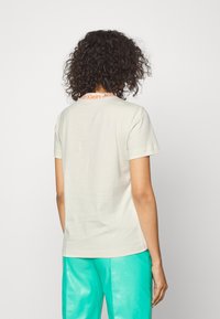 Woman with curly hair wearing a light beige T-shirt with orange collar text "Calvin Klein Jeans" and turquoise pants, shown from behind.