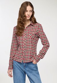 Giovane donna con lunghe onde nei capelli, che indossa una camicia a scacchi rossa e beige e jeans blu, in piedi con una mano in tasca, che guarda a destra.