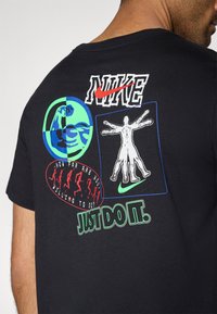 Μαύρο T-shirt της Nike με γραφικό σχέδιο που περιλαμβάνει διάφορα έγχρωμα τυπώματα, όπω ς έναν μυώδη βραχίονα, μια ανθρώπινη φιγούρα και τη φράση «Απλώς κάντο».