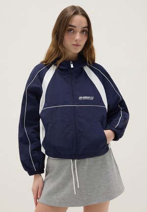 Young woman wearing a navy and white zip-up sports jacket with "Los Angeles Sport Club" text and a grey skirt, standing with one hand in pocket.