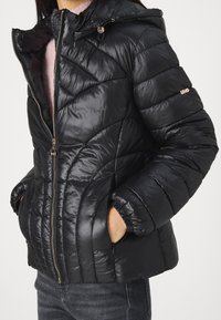 Giubbotto imbottito nero con finitura lucida, cappuccio e design a cuciture diagonali. Presenta chiusura con zip e tasche laterali per funzionalità.