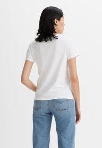 Weißes kurzärmliges T-Shirt mit Rundhalsausschnitt, getragen von einer Person, die hellblaue hochgeschnittene Jeans mit Gesäßtaschen trägt.