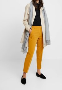 Beżowy trencz na czarnym topie, połączony z musztardowymi spodniami. Szary szal z frędzlami i czarne loafersy dopełniają stylizację.