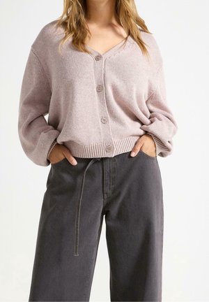 Femme portant un cardigan en maille mauve clair boutonné avec manches longues et un pantalon large taille haute foncé, mains dans les poches, sur fond uni.