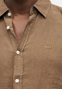 Gros plan d'une personne portant une chemise marron à boutons blancs avec le mot BOSS brodé sur la poitrine.