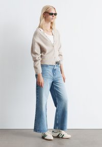 Hellbeige Strickjacke, lockere Passform, mit Knopfleiste; kombiniert mit weit geschnittenen, hellblauen Jeans und weißen Sneakers mit grünen Streifen.