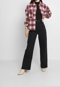 Koszula z flaneli w czerwono-kremową kratę, czarne spodnie z wysokim stanem oraz beżowe buty na grubym obcasie. Koszula ma przednie guziki i kieszenie.