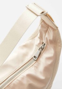 Borsa in tessuto beige con forma arrotondata, dotata di una cerniera argentata e di una tracolla in crema abbinata. Texture morbida con dettagli di cucitura visibili.