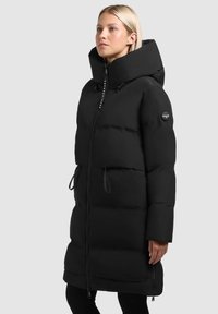 Giacca a vento nera con colletto alto, zip frontale, cordini e una patch con logo. Presenta un design trapuntato e una silhouette oversize.