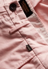 Gros plan sur un pantalon rose clair avec passant de ceinture, bouton noir et petite étiquette noire de la marque Superdry près de la taille.