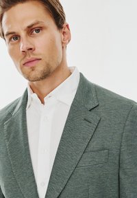Dopasowana marynarka z teksturowanej zielonej tkaniny, z wykładanym kołnierzem, pojedynczą kieszenią na piersi, noszona na białej koszuli z guzikami.